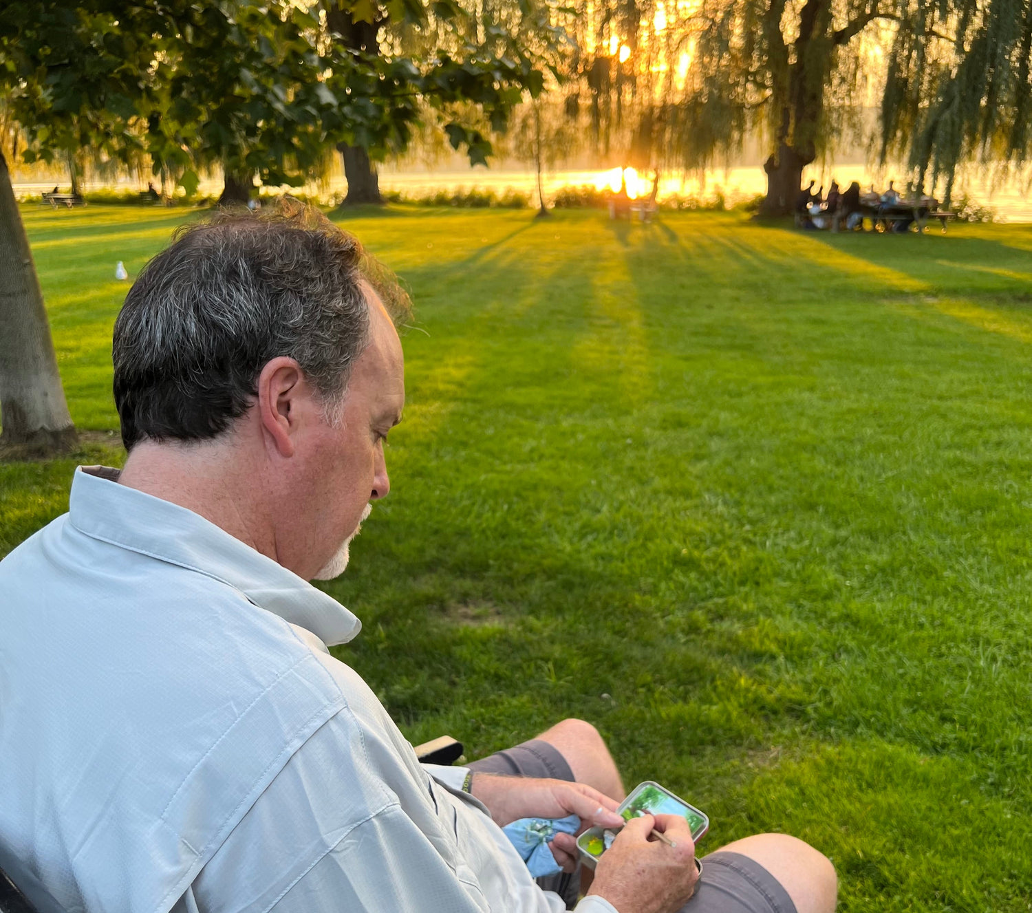 Man sitting on grass in a park painting a scene in an Altoid box with sunset and trees in the background